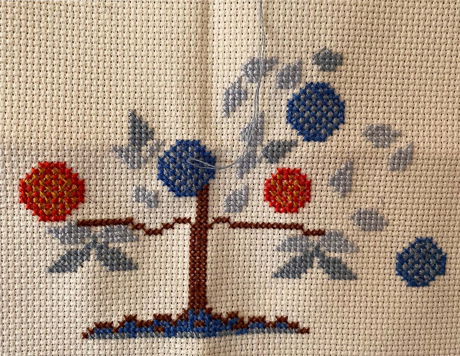 Close-up of an in-progress cross stitch work: a tree of life, with the leaves in blues and orange. A need with blue thread is inserted into the middle of the fabric.